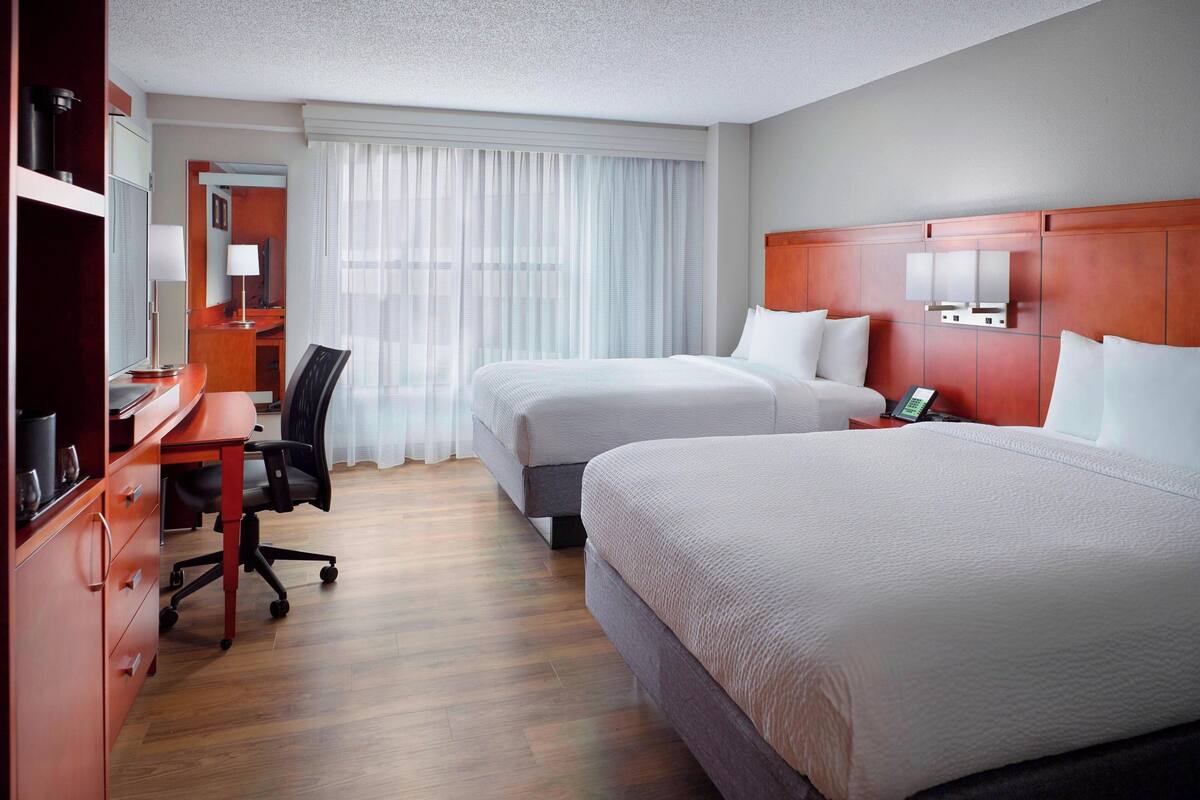 A hotel room with two beds, a desk with a chair, a TV, and a bathroom visible through a door.