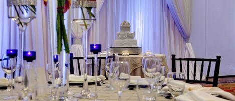 A formal dining table set with wine glasses, a cake centerpiece, and white tablecloths.