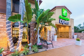 A Holiday Inn Express building with a stone facade, large tropical plants, and a bench outside. - Holiday Inn Express San Diego - SeaWorld Area by IHG (San Diego)