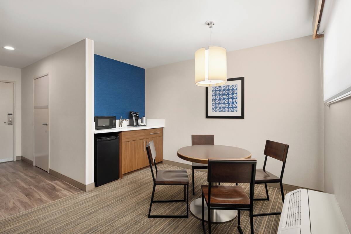A compact hotel room with a kitchenette, a dining area with a round table and four chairs, a microwave, a coffee maker, and a framed artwork on the wall.