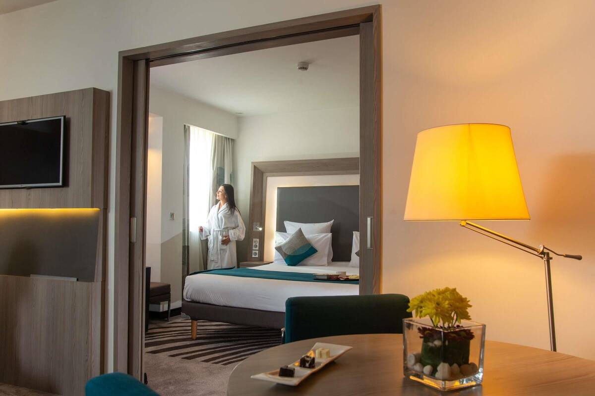 Una habitación de hotel con cama, televisión, lámpara, mesa con un florero y una silla.