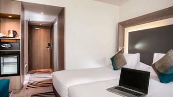 Ein Hotelzimmer mit einem Bett, einem Laptop, einem Schreibtisch mit Kaffeemaschine und einer Tür zu einem anderen Raum.