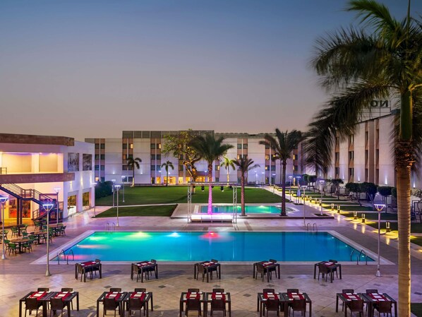 A hotel pool area with multiple dining tables and chairs, a large pool, and palm trees.