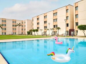 A swimming pool with inflatable unicorn and flamingo floats, surrounded by a building with multiple floors and a flat roof.