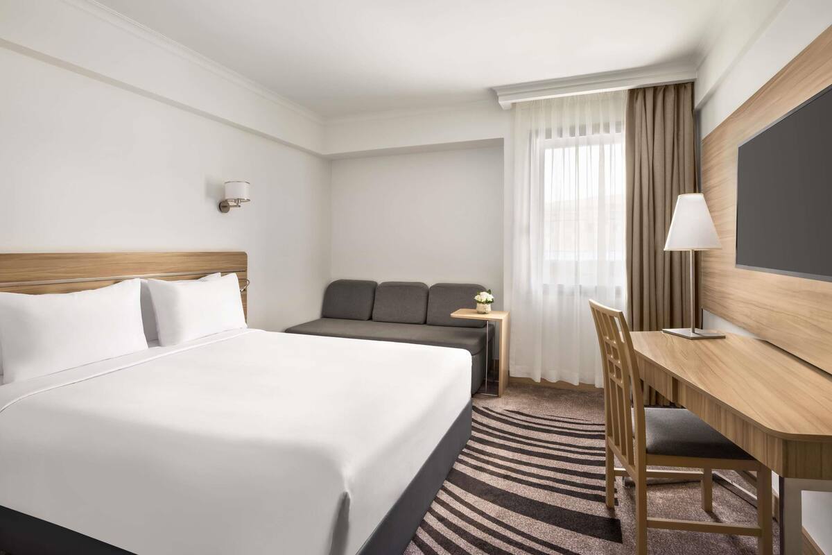 Habitación de hotel con cama, escritorio, silla, sofá, televisor y ventana con cortinas.