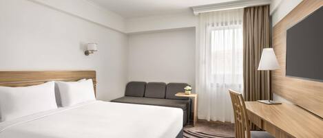 A hotel room with a bed, a desk, a chair, a sofa, a TV, and a window with curtains.