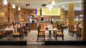 A restaurant with a modern interior, featuring a long counter with a chef preparing food, and multiple dining tables with chairs. - Novotel Cairo Airport (Cairo)