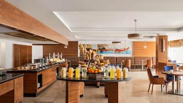 Un hall d’hôtel moderne doté d’un espace de restauration central où se trouve un buffet proposant une variété de plats et de boissons. On y trouve également des coins salon avec des canapés et des tables, ainsi qu’un grand tableau abstrait accroché au mur.