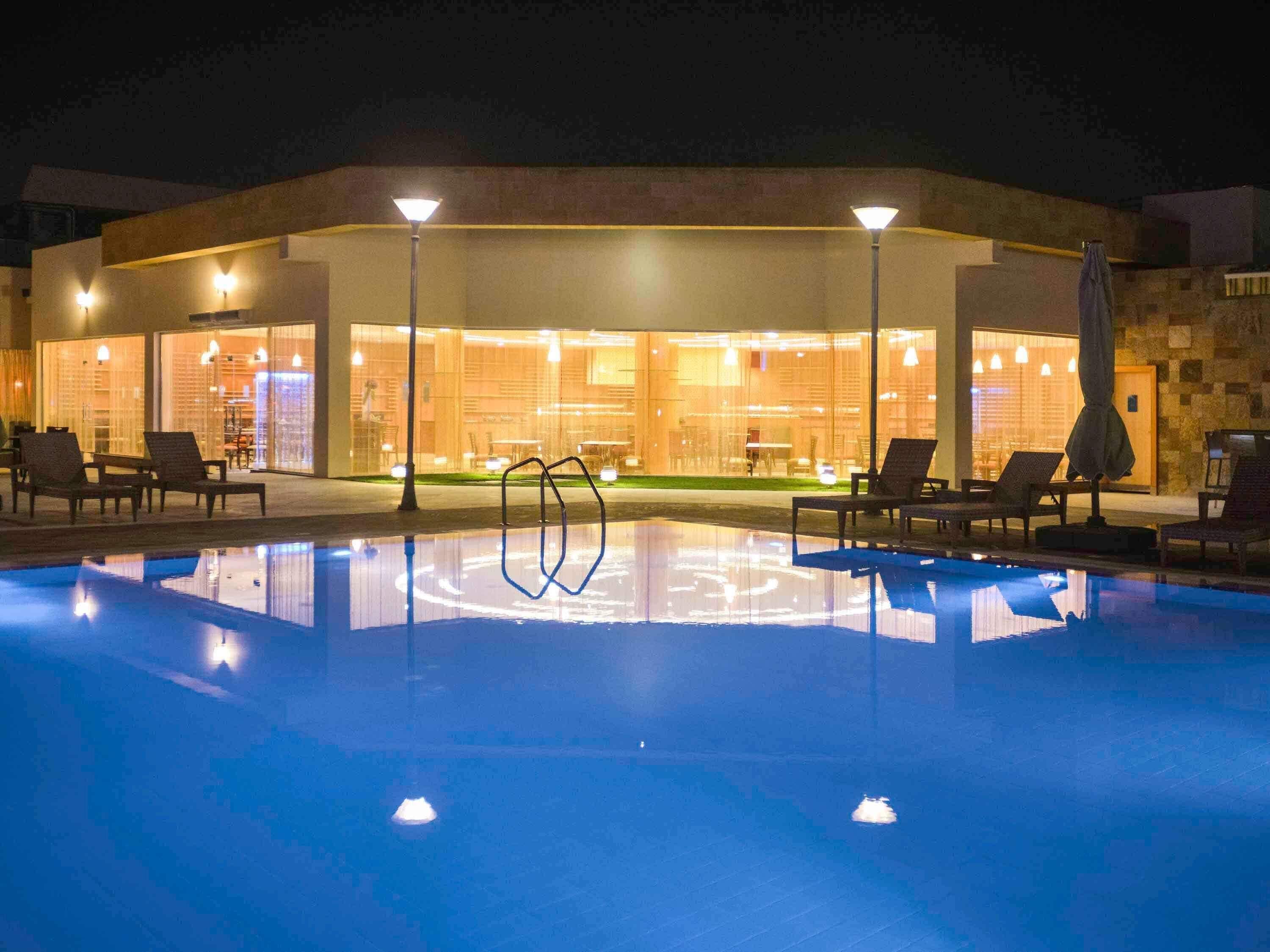 Piscina com espreguiçadeiras e guarda-sóis, restaurante com paredes de vidro e fachada de pedra.