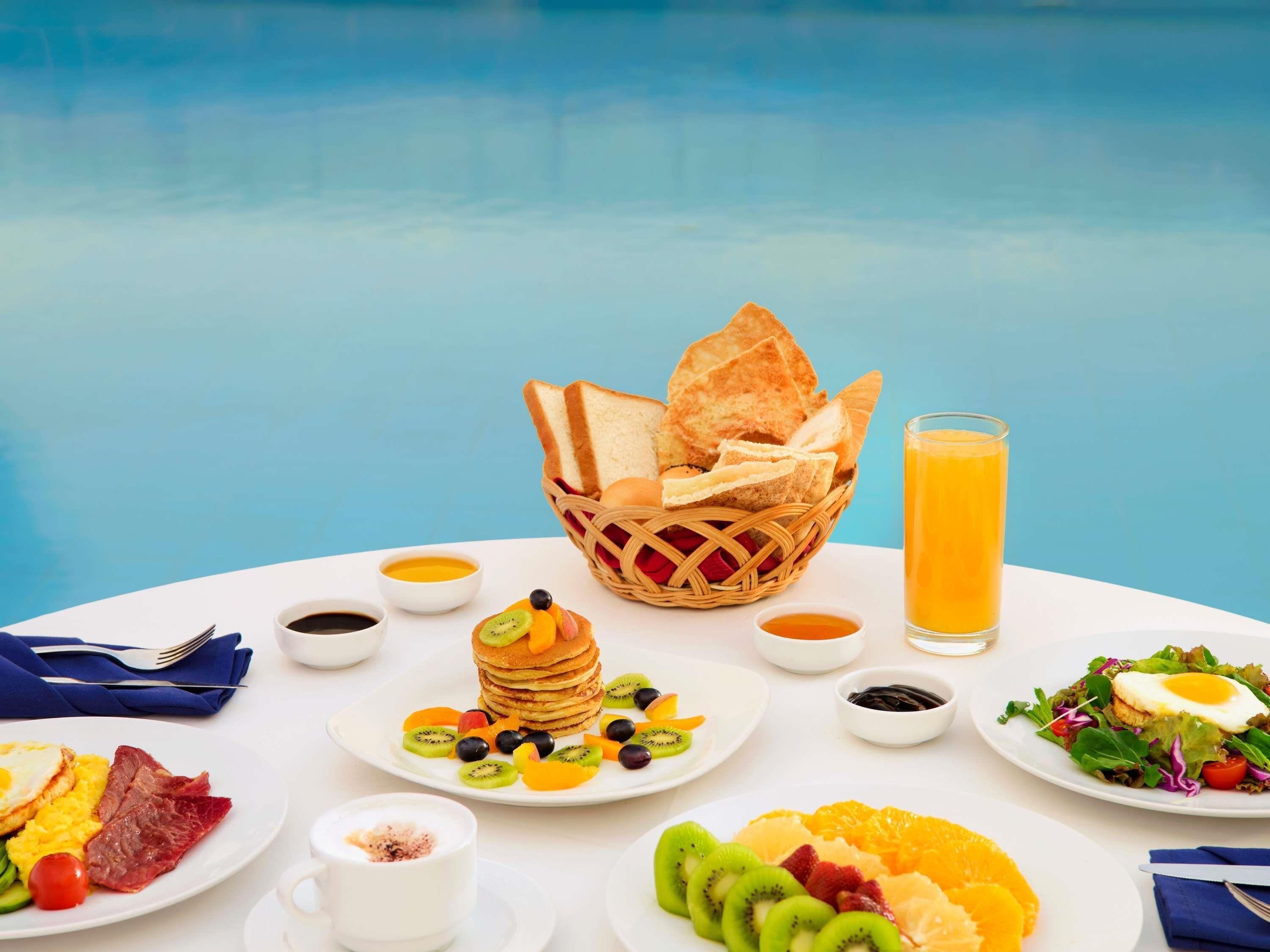 A breakfast table with various dishes including toast, pancakes, eggs, and fruit, set against a poolside view.