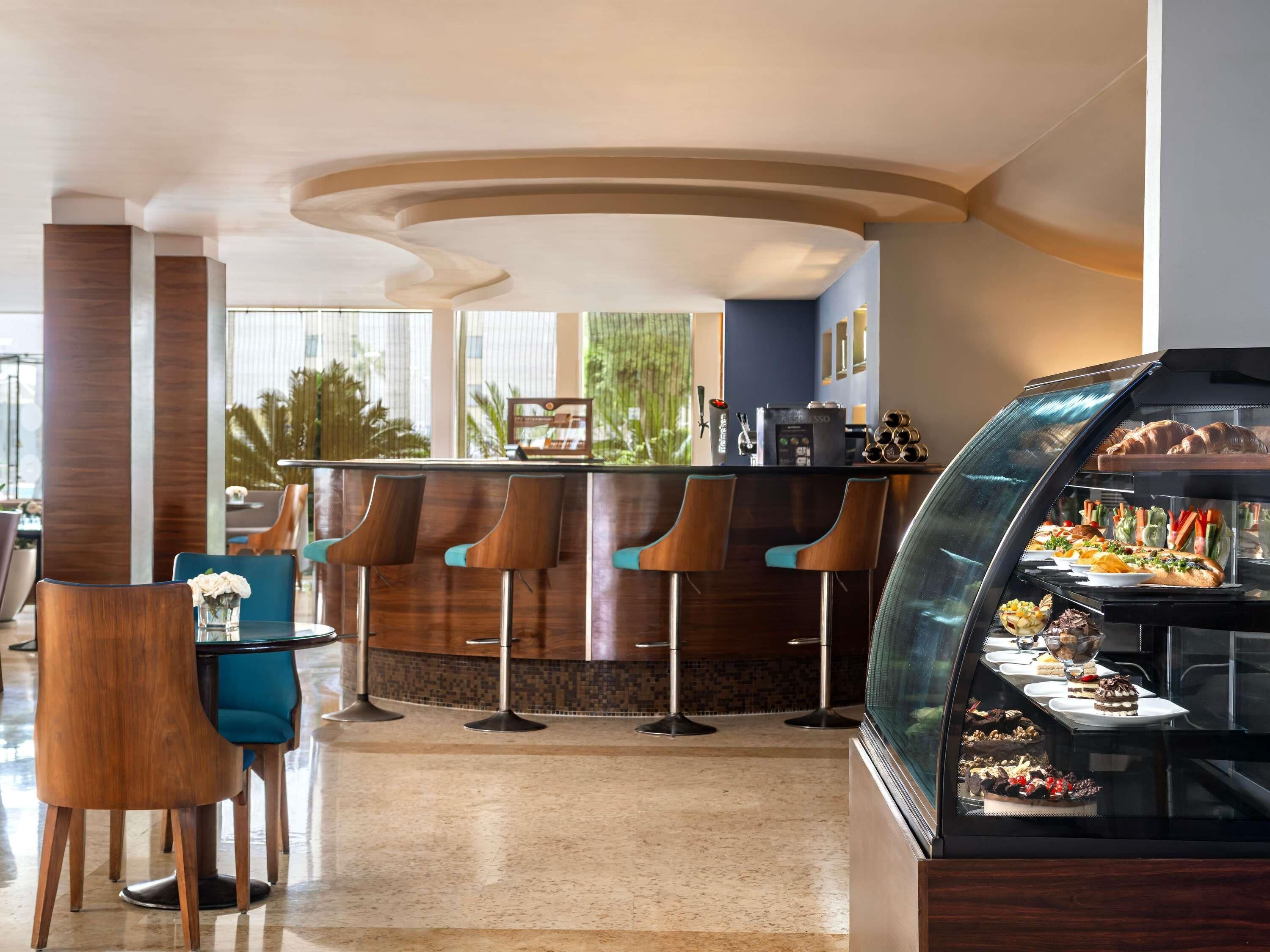 A modern bar area with a curved ceiling, a glass display case with desserts, and a seating area with bar stools and a table.