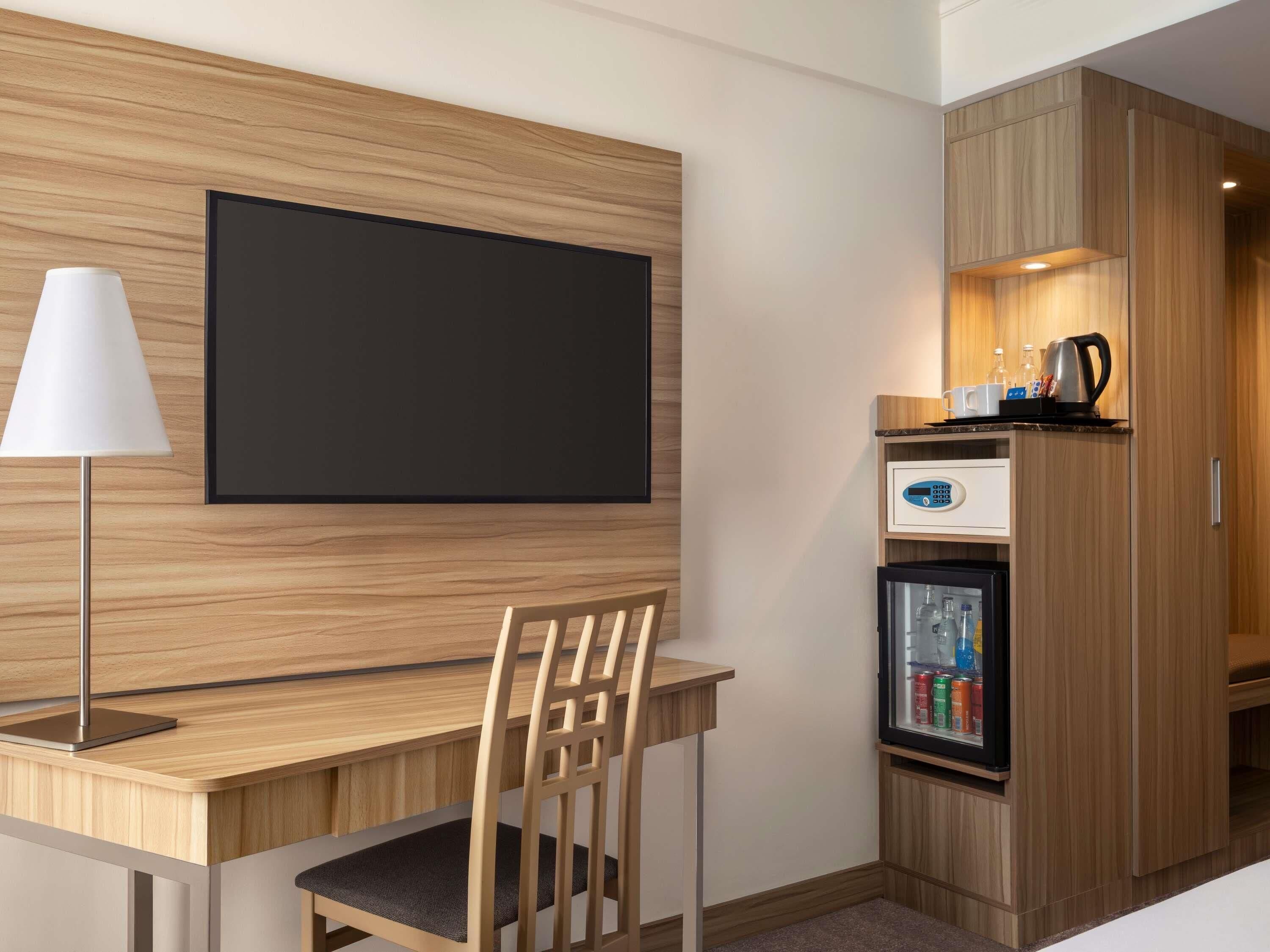A hotel room with a wooden desk, a chair, a television mounted on the wall, a refrigerator with beverages, and a coffee maker.