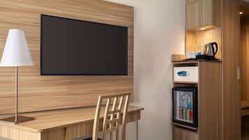A hotel room with a wooden desk, a chair, a television mounted on the wall, a refrigerator with beverages, and a coffee maker.