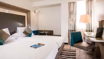 A hotel room with two beds, a desk with a laptop, a chair, a small table with a lamp, and a window with curtains.