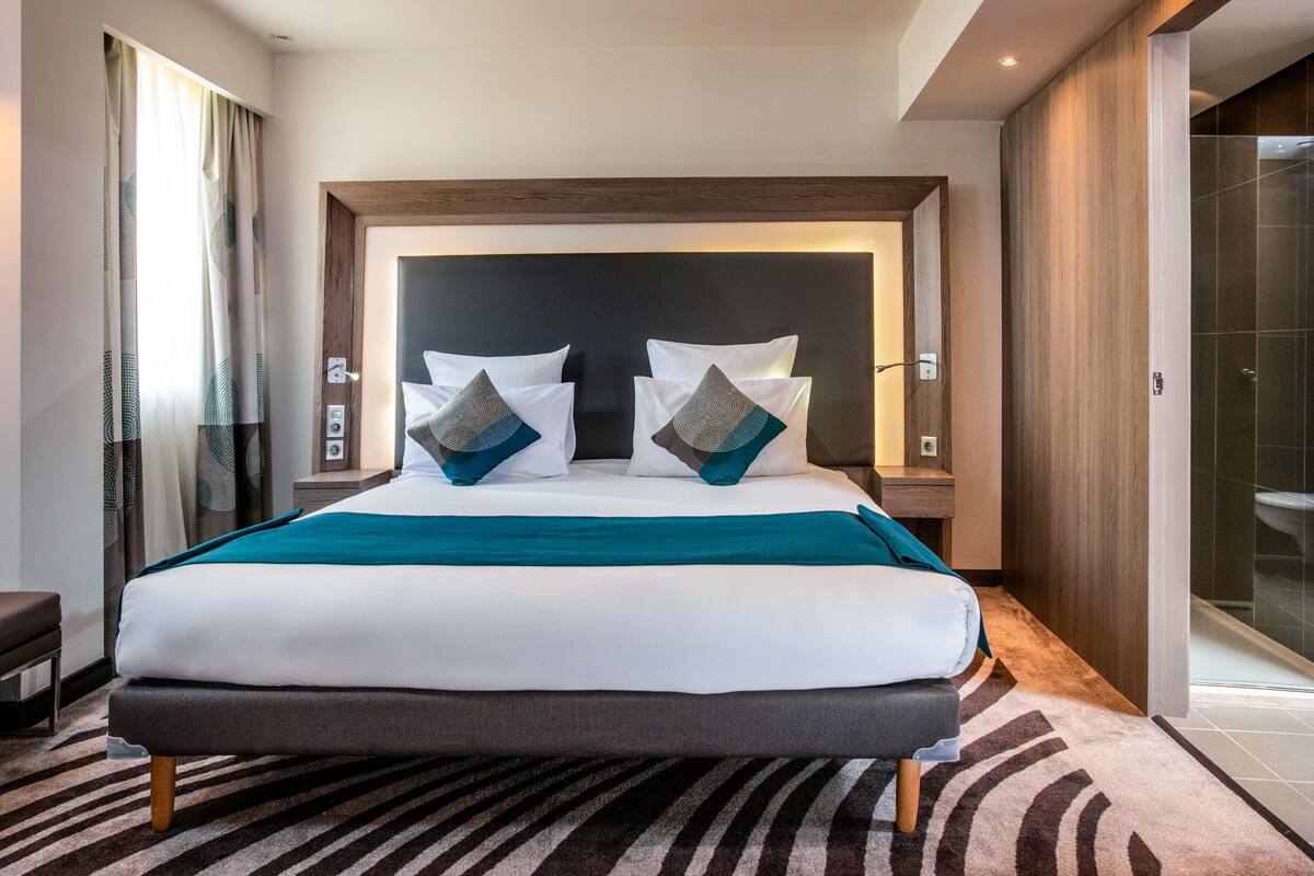 A modern hotel room with a large bed, a headboard, bedside tables, a bench, and a view of the bathroom.
