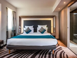 A modern hotel room with a large bed, a headboard, bedside tables, a bench, and a view of the bathroom.