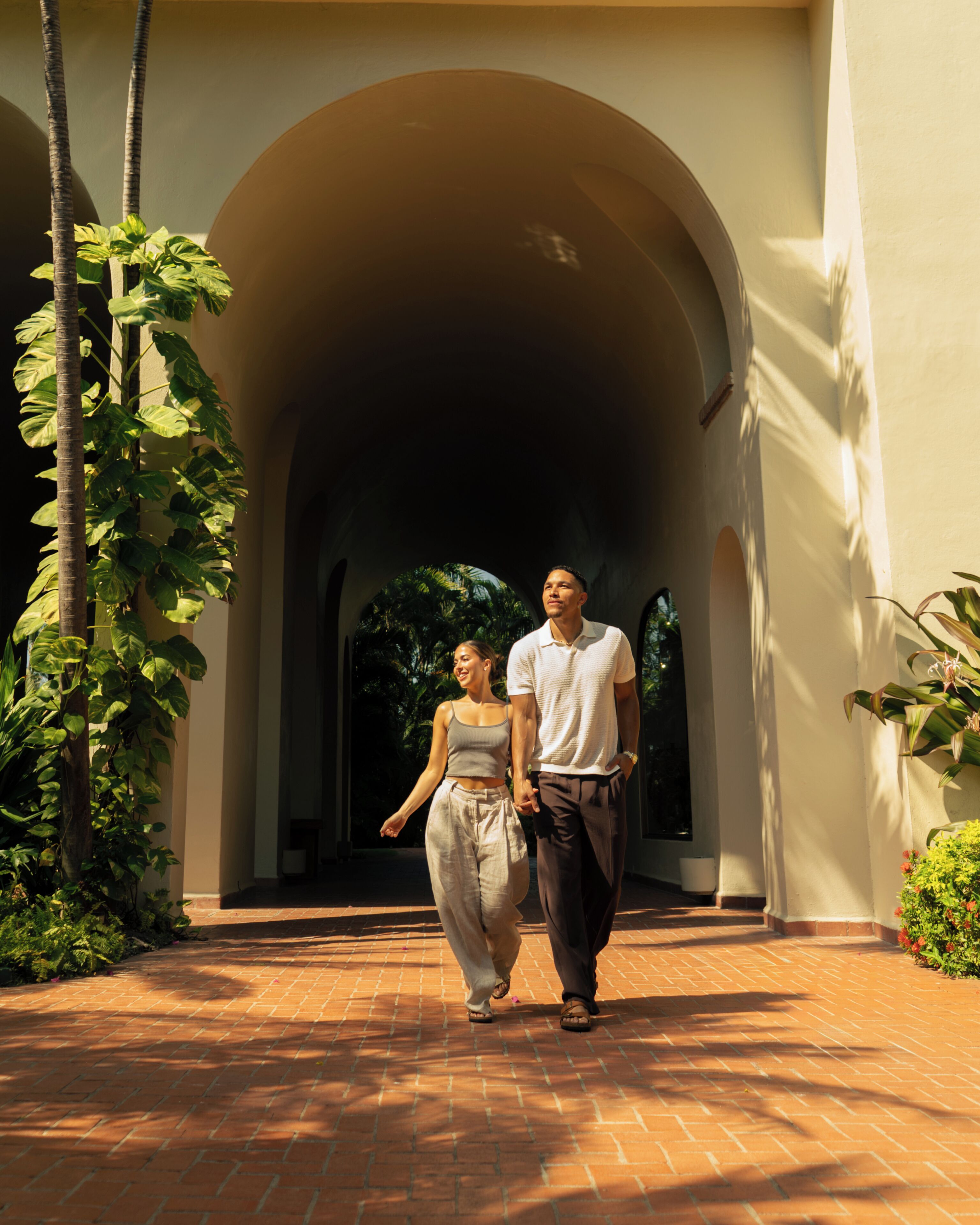 A couple walking through an arched corridor with a brick floor and greenery on the sides.