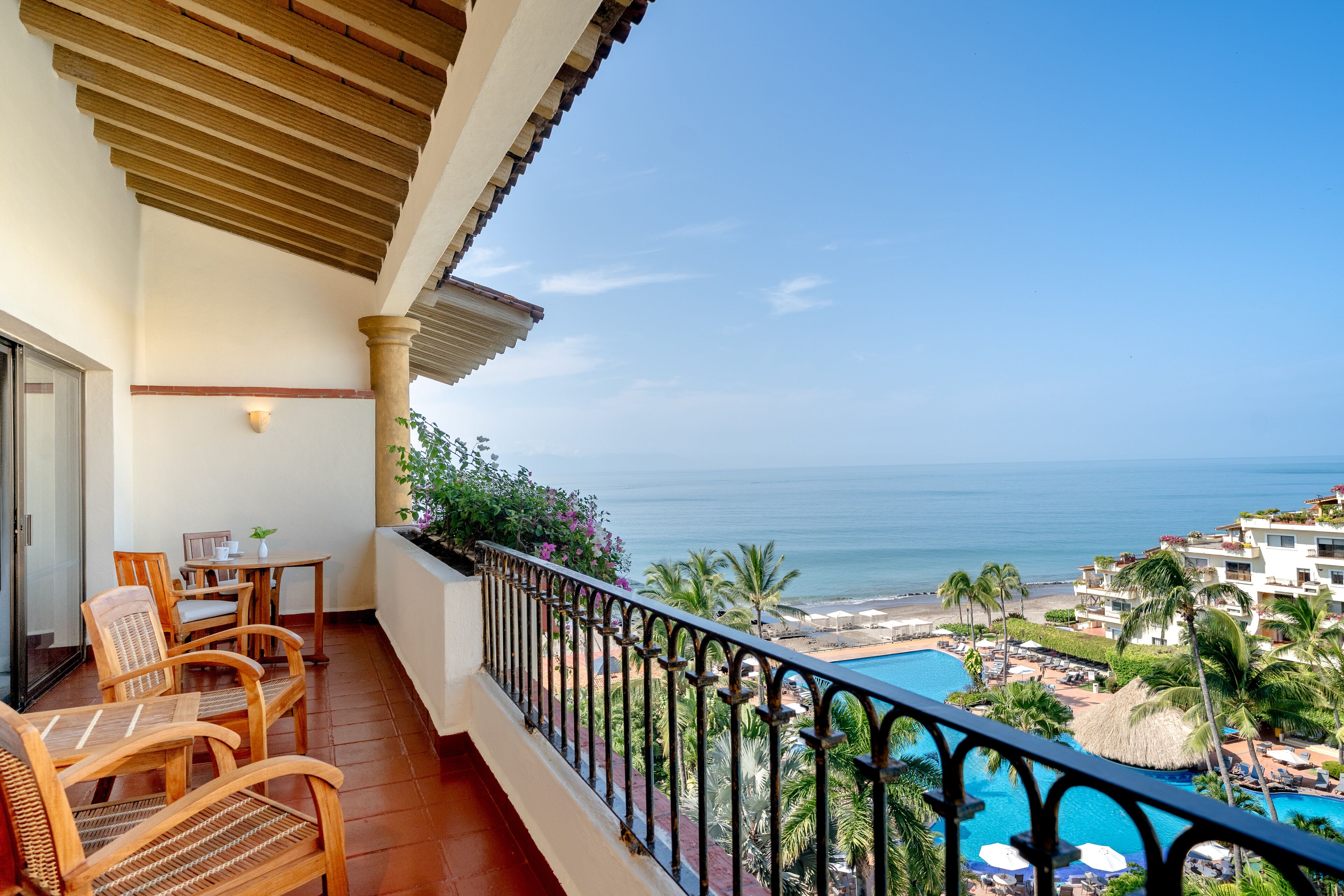 A balcony with wooden chairs, a table, and a view of the ocean and a swimming pool.