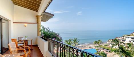 A balcony with wooden chairs, a table, and a view of the ocean and a swimming pool.