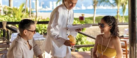 A person serving a drink to two seated individuals at an outdoor table with a view of the ocean and palm trees.