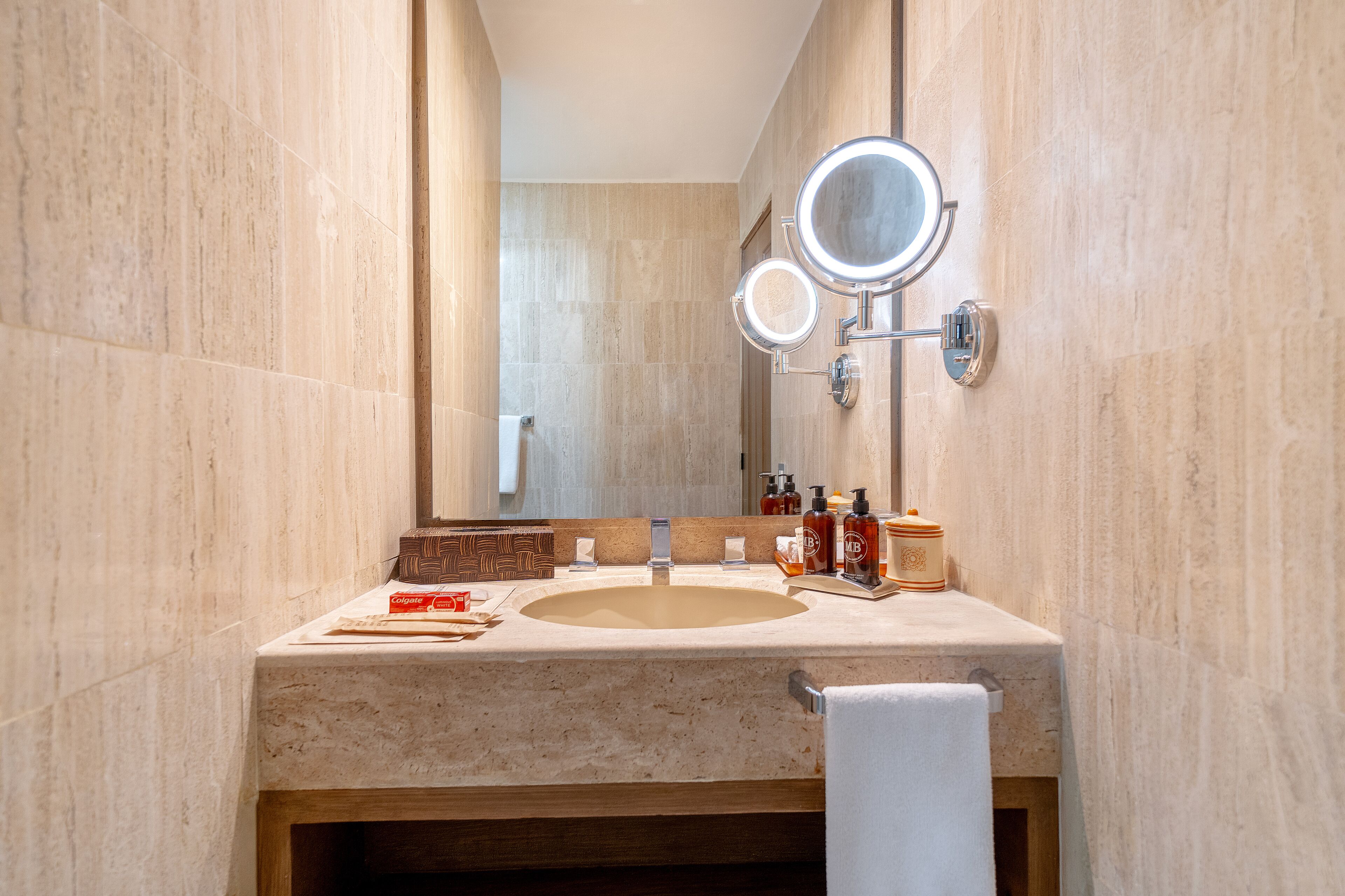 Un baño con un lavabo de mármol color beige, dos espejos redondos con iluminación, artículos de aseo y un perchero para toallas.