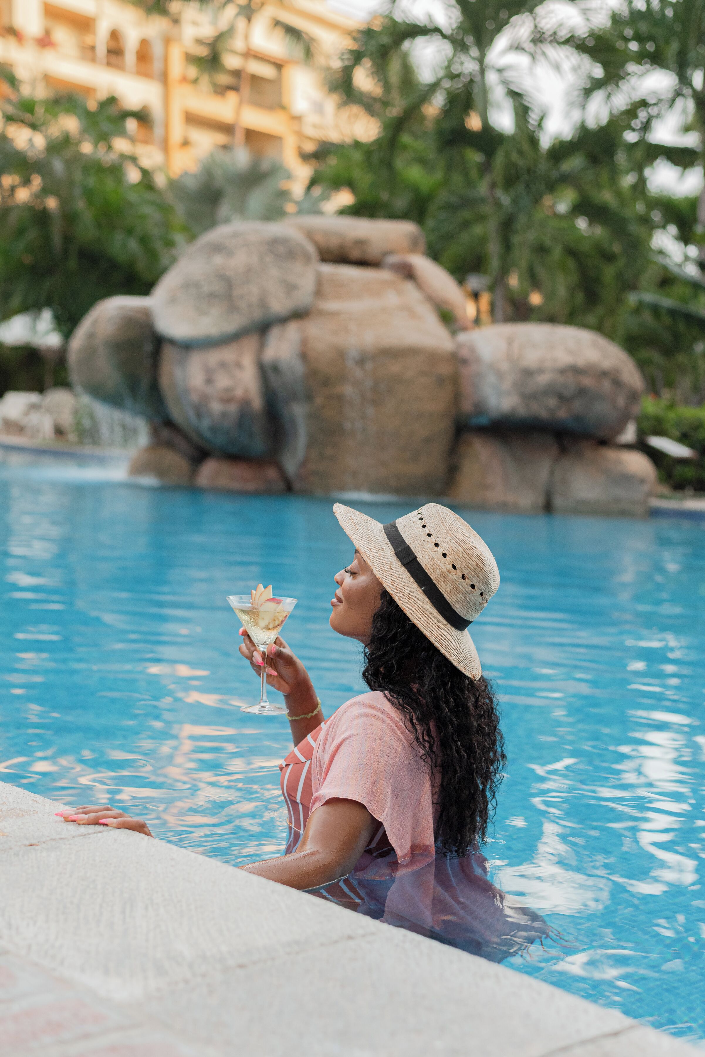 Une personne assise au bord d’une piscine, un verre à la main, entourée de gros rochers et de verdure.