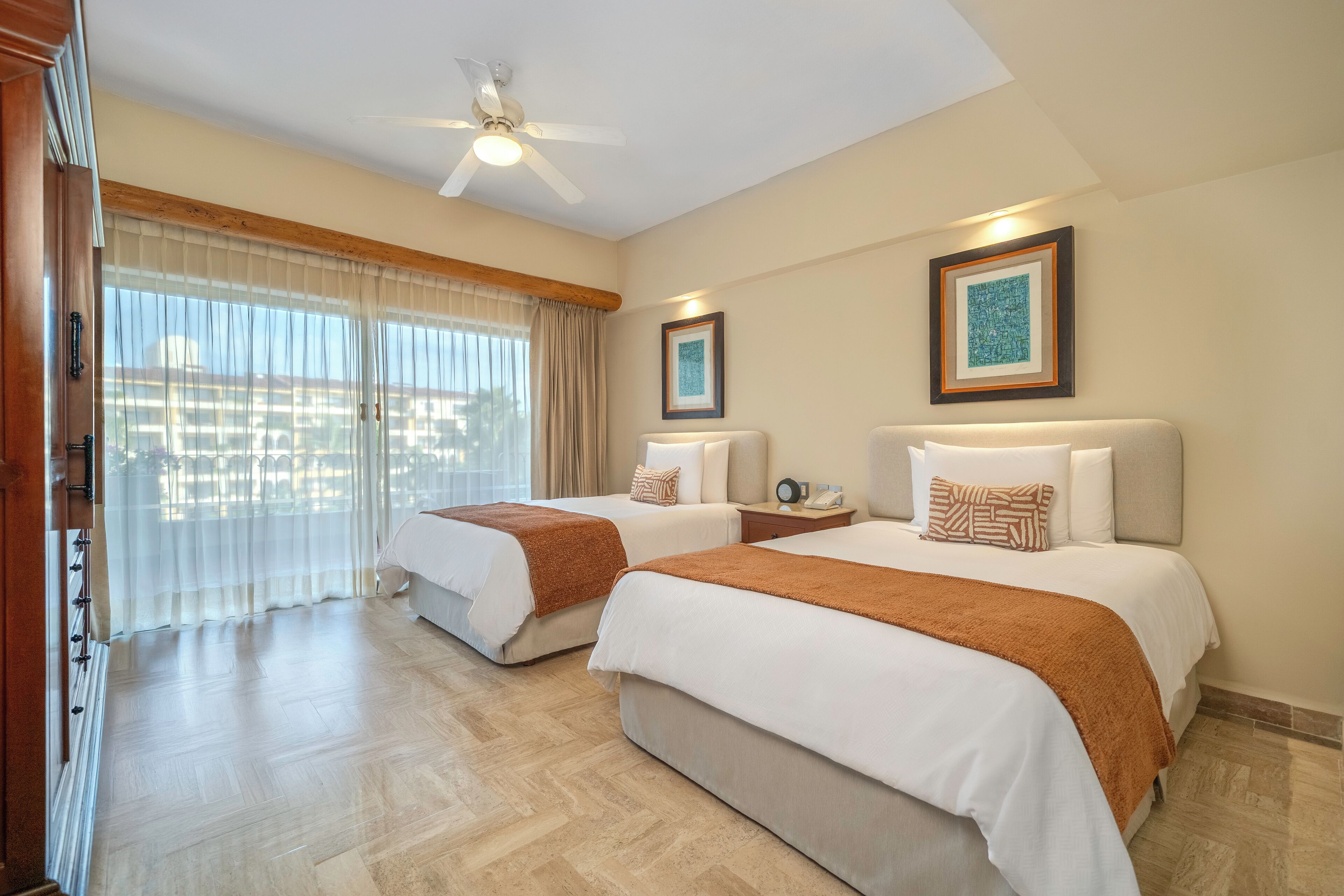 A hotel room with two beds, a ceiling fan, and large windows with sheer curtains.