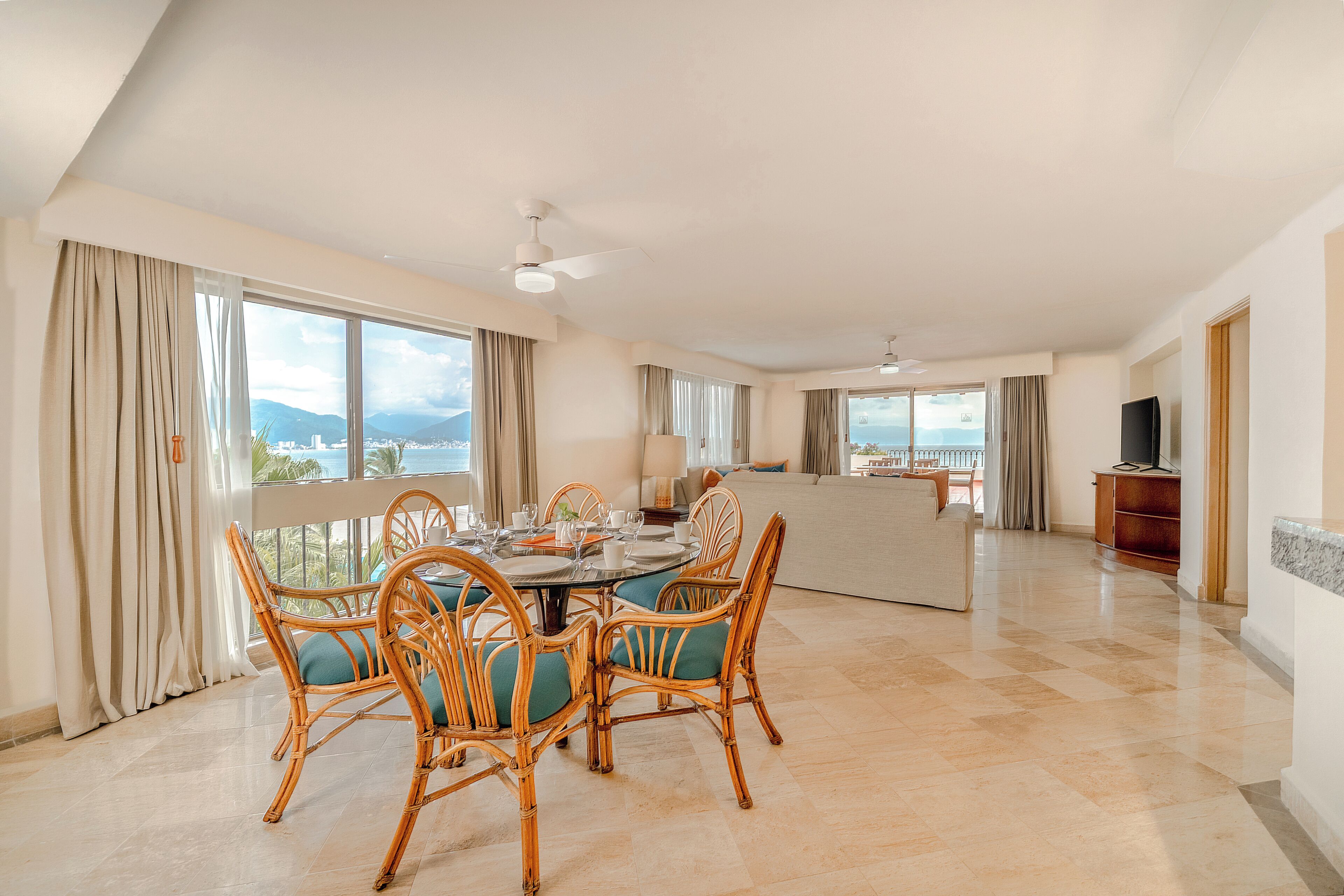 A spacious living area with a dining table set for four, surrounded by wicker chairs with green cushions. A large window offers a view of the city and mountains.