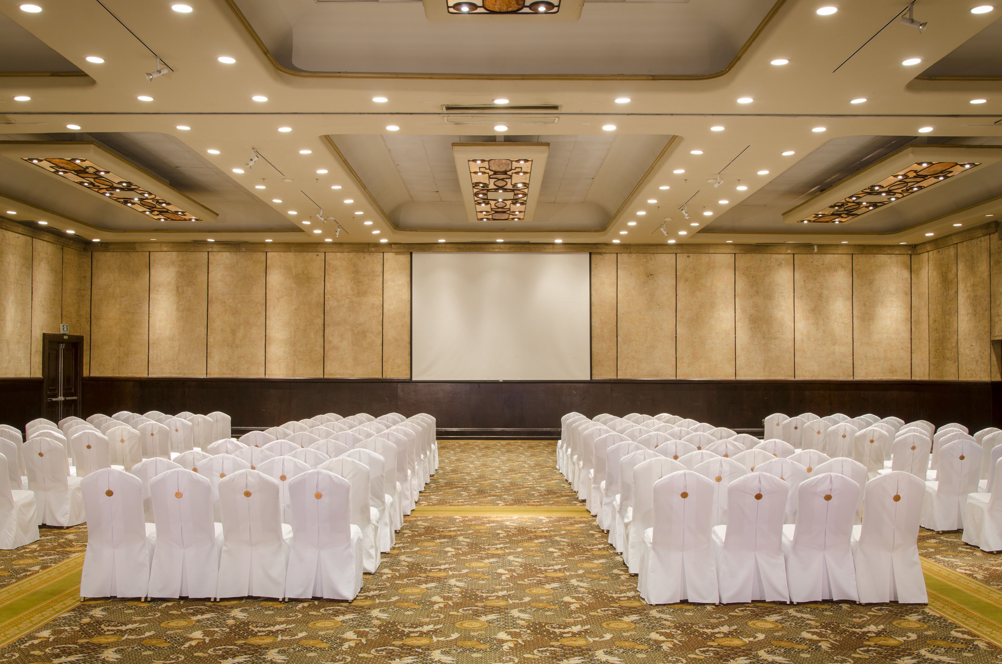 Un salón de conferencias con filas de sillas blancas, suelo alfombrado y una pantalla grande al frente.