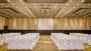 Un salón de conferencias con filas de sillas blancas, suelo alfombrado y una pantalla grande al frente.