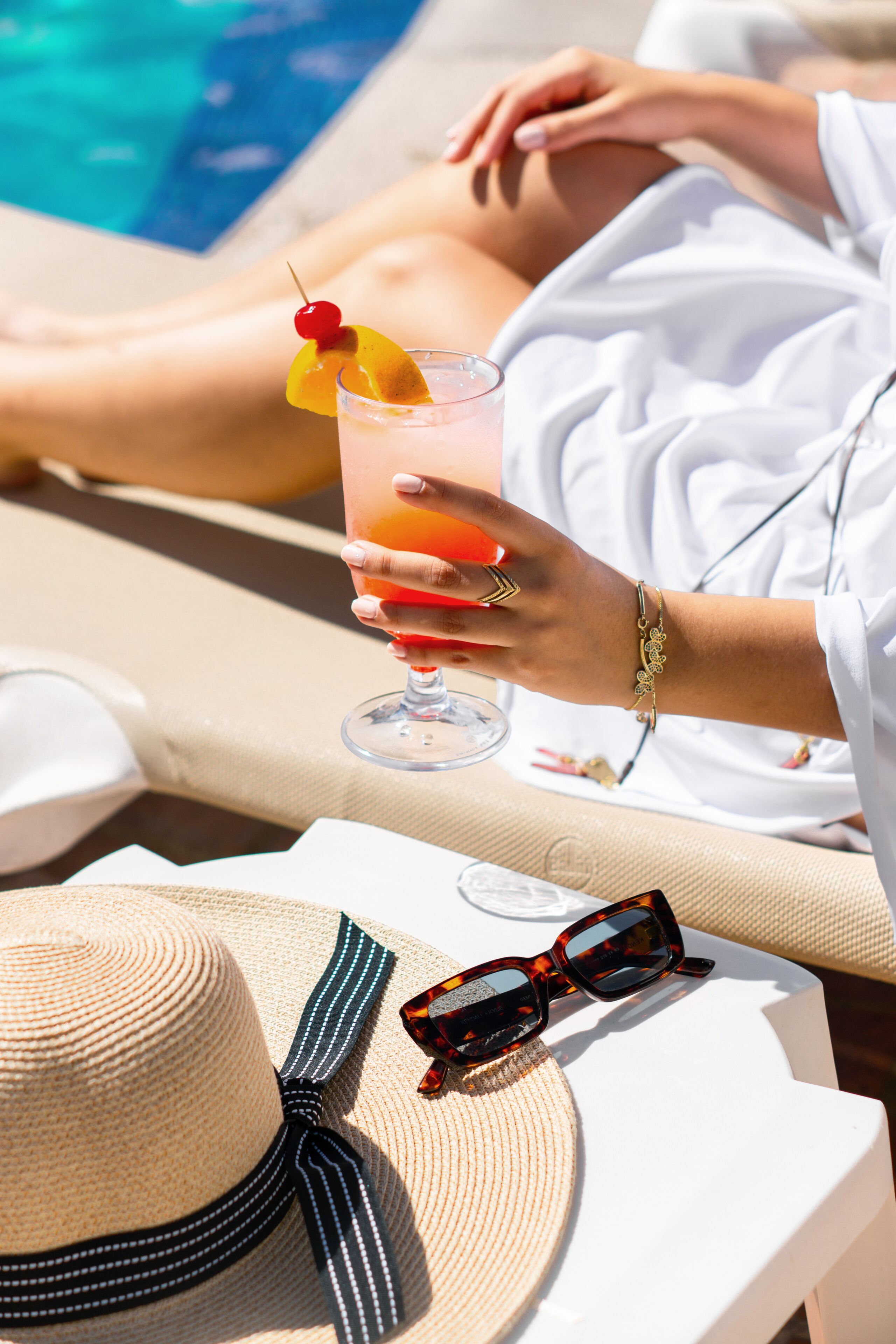 A poolside setting with a person relaxing, a drink with a cherry garnish, a straw hat, and sunglasses on a table.