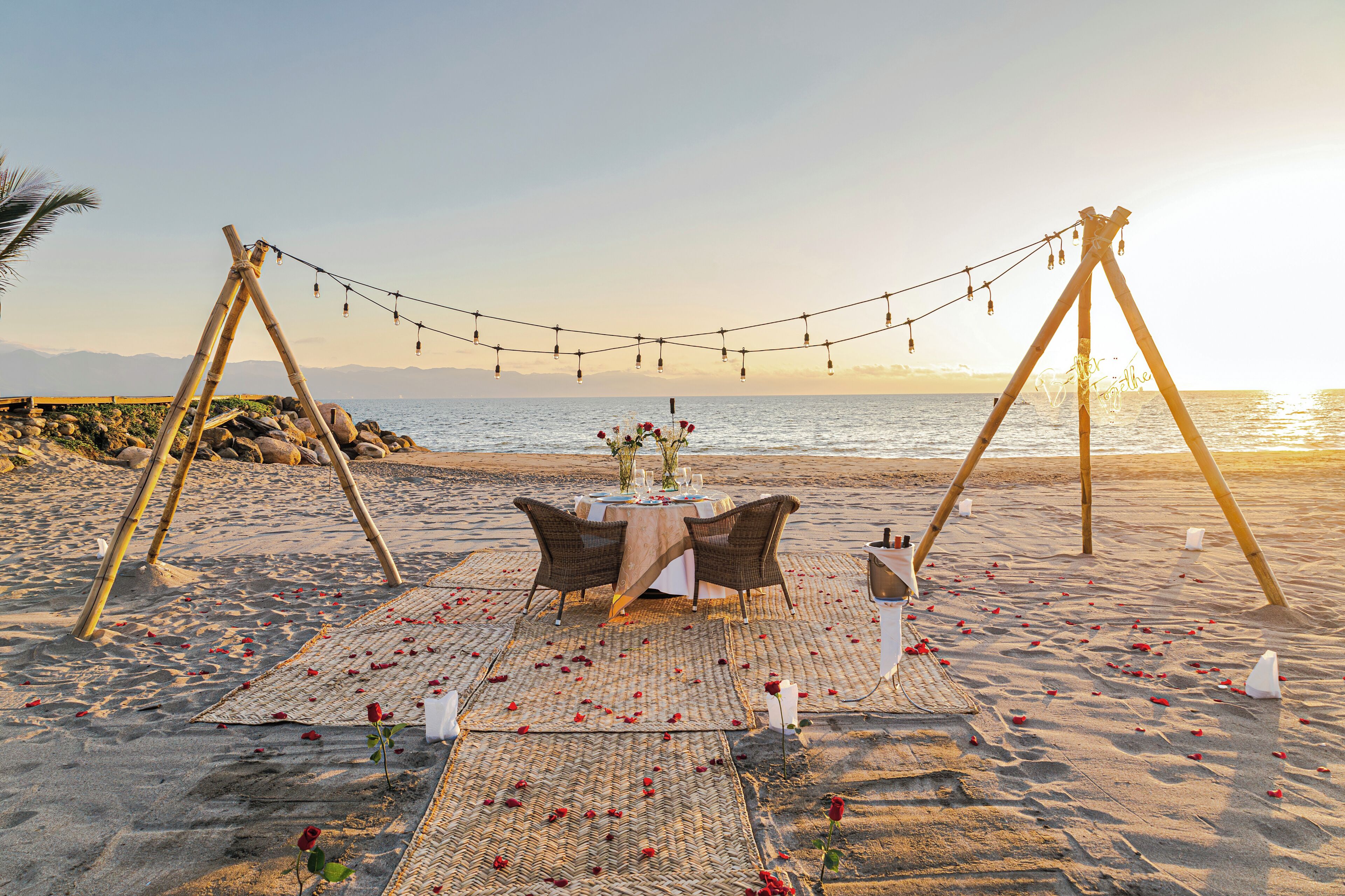 A beach setting with a table set for two, surrounded by rose petals and a string of lights.