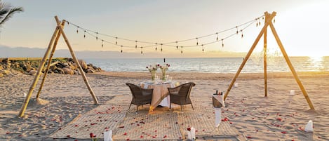 A beach setting with a table set for two, surrounded by rose petals and a string of lights.
