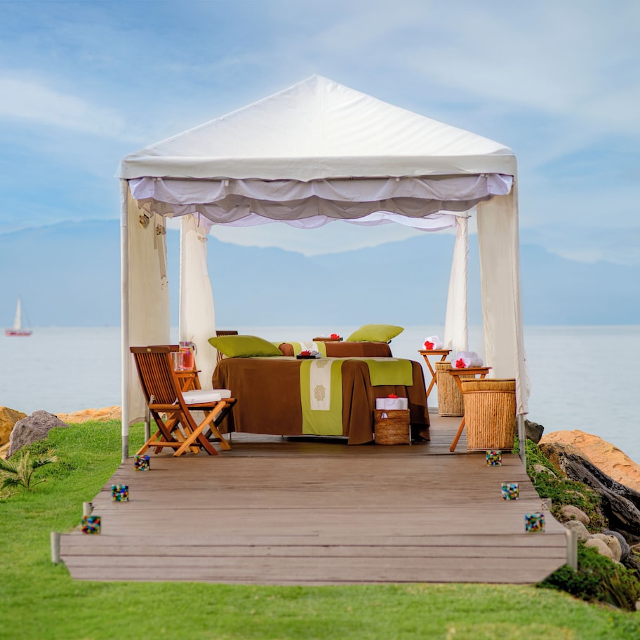 A tented outdoor area with a bed, chairs, and a table, overlooking a body of water with rocks and a distant sailboat.