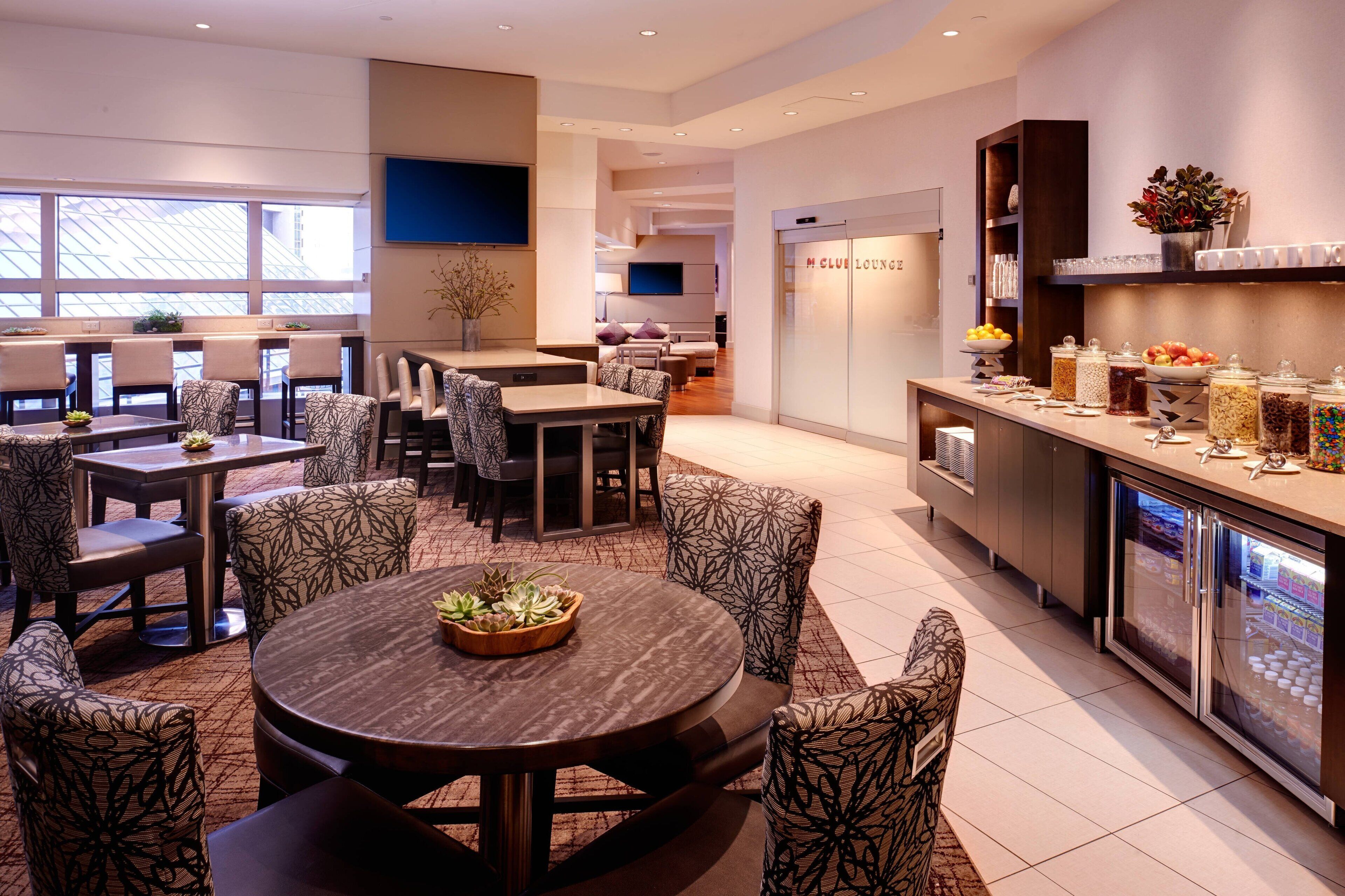 A modern hotel lobby with a dining area, a lounge, and a kitchen counter with food dispensers.