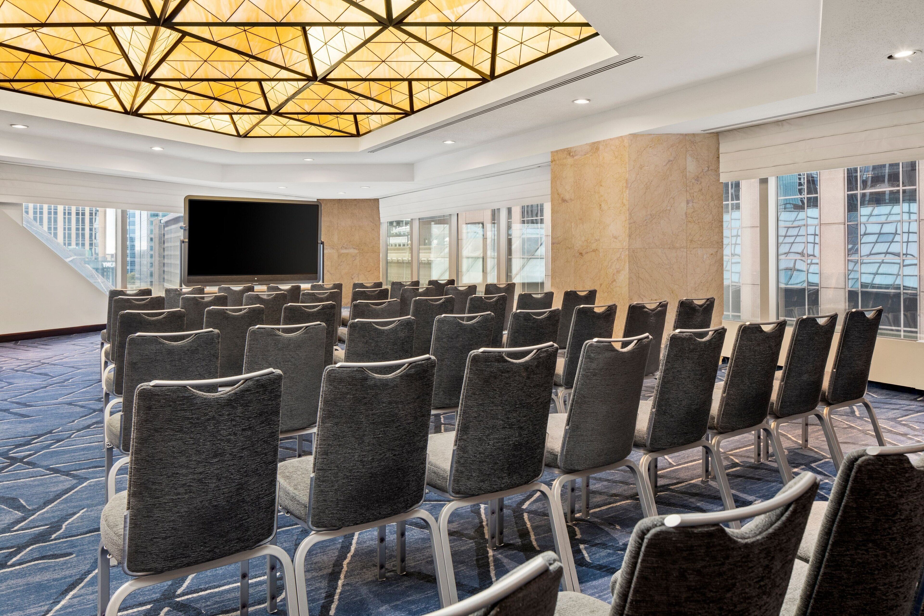 Una sala conferenze con file di sedie grigie, un grande schermo e un design geometrico del soffitto.