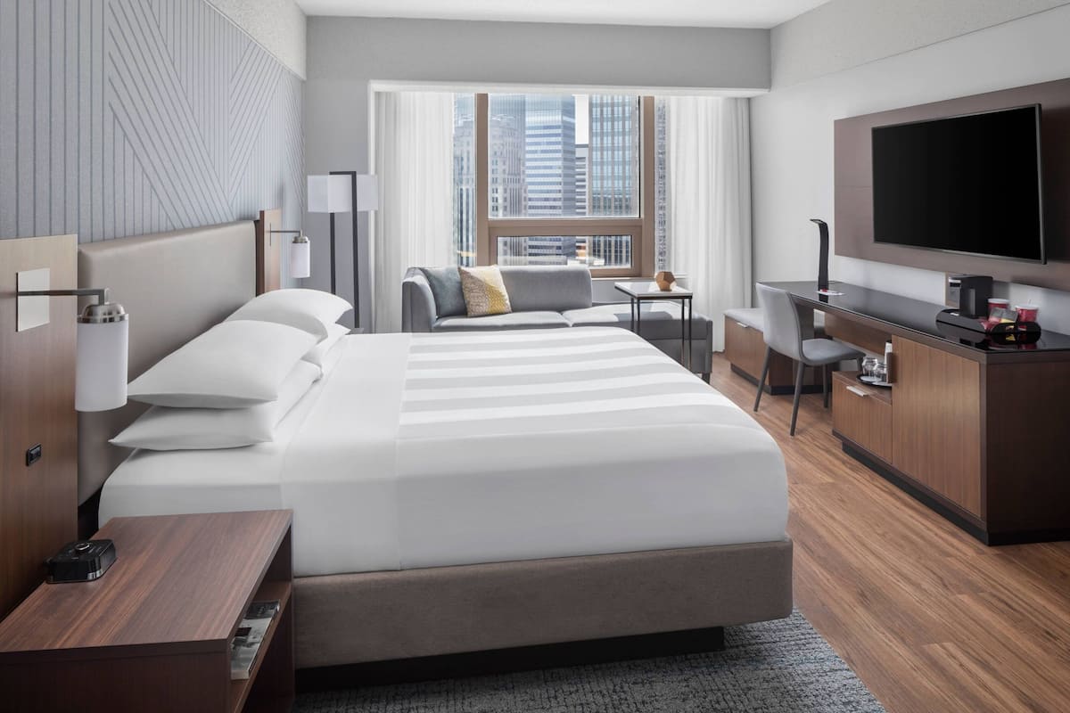 A modern hotel room with a large bed, a desk, a flat-screen TV, and a city view.