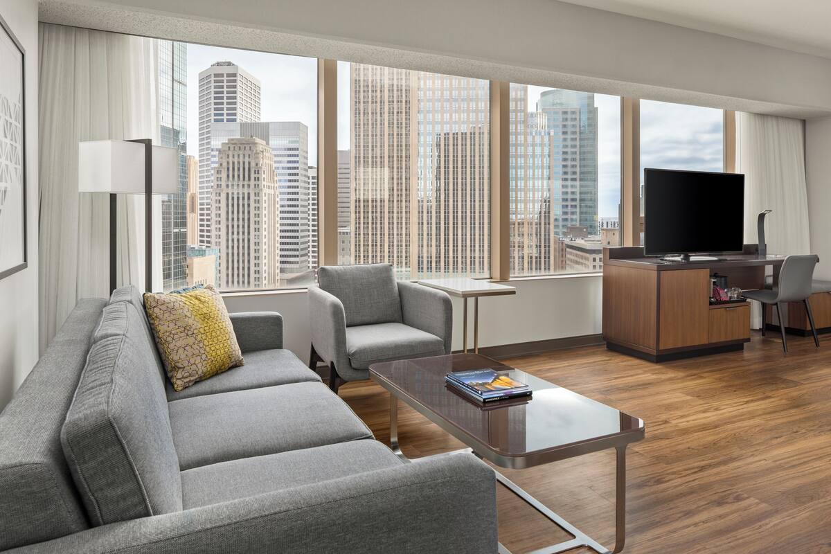 A modern hotel room with a grey sofa, a small table, a TV, and a city view.