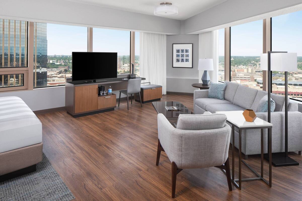 A modern hotel room with a large bed, a sofa, a desk with a flat-screen TV, and a city view.