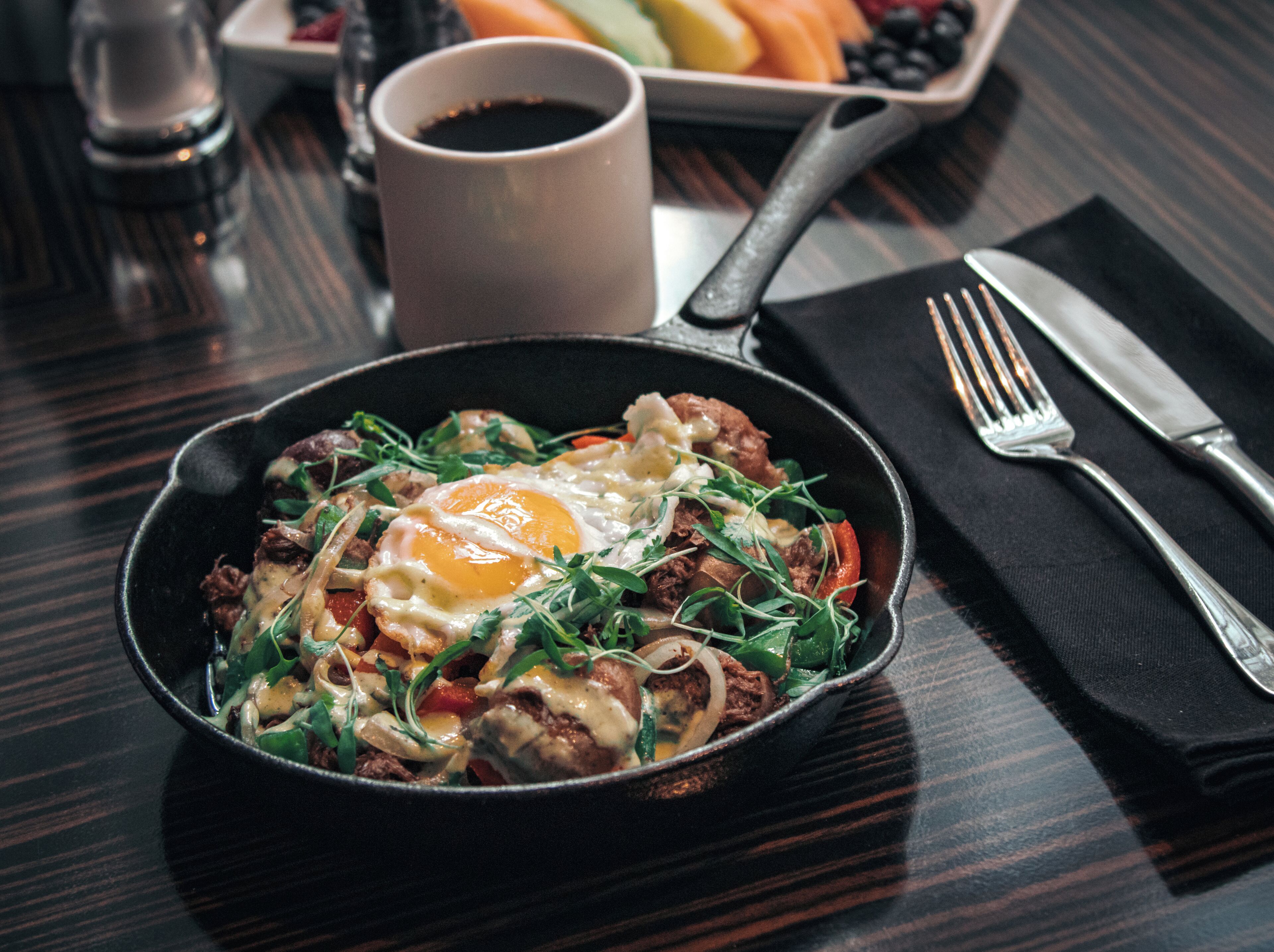 A breakfast dish in a skillet, a cup of coffee, and a plate of fruit on a table.