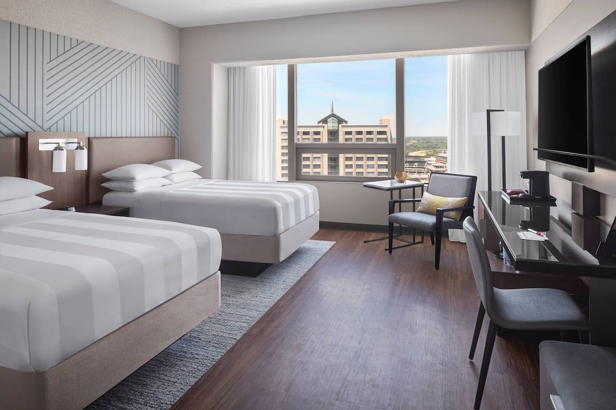 A hotel room with two beds, a desk with a chair, a TV, and a large window with a city view.