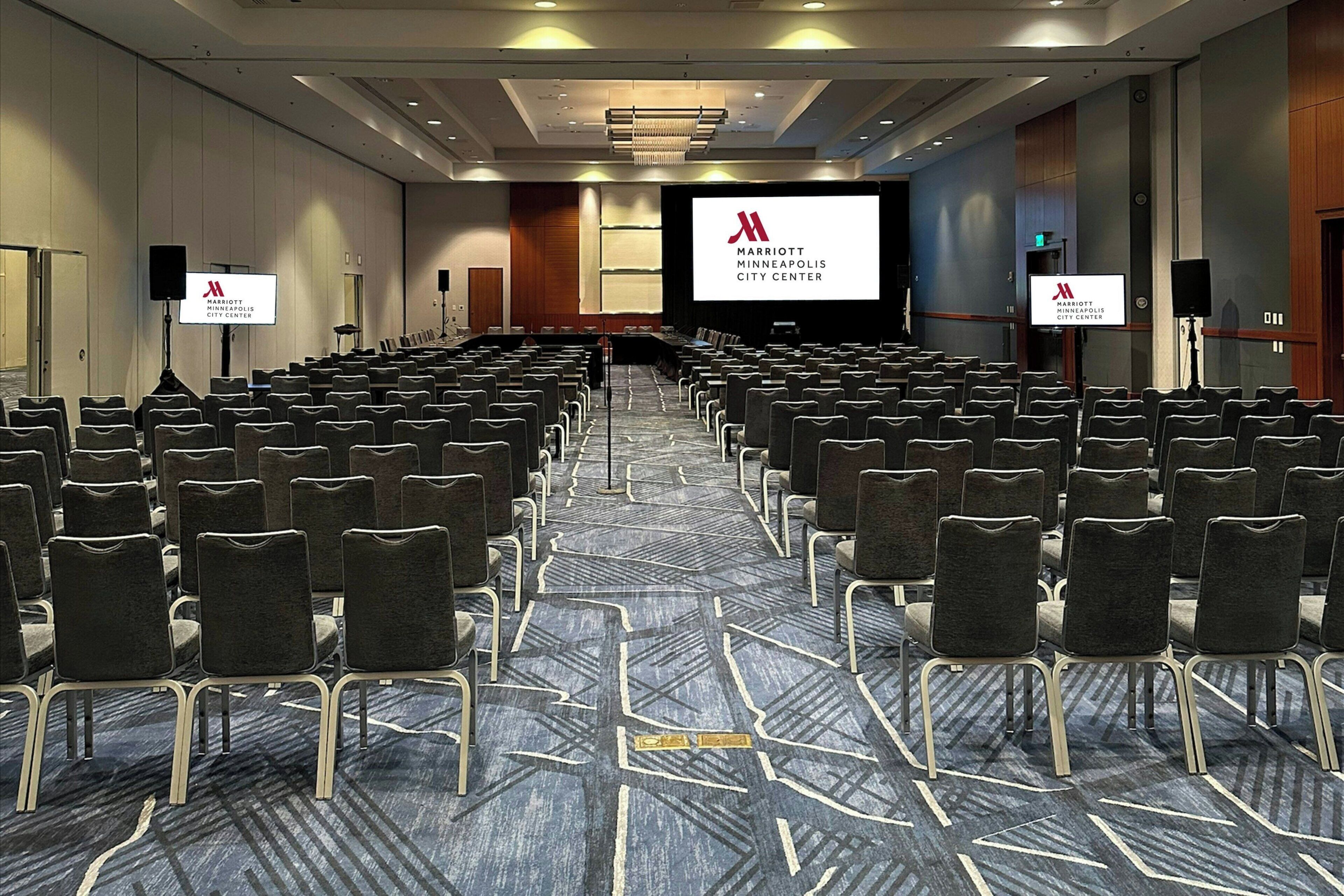 Un salón de conferencias con filas de sillas, una pantalla grande mostrando el logotipo de Marriott Minneapolis City Center y un piso alfombrado.