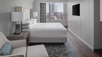 A modern hotel room with a large bed, a sofa, a desk with a lamp, and a TV on the wall.