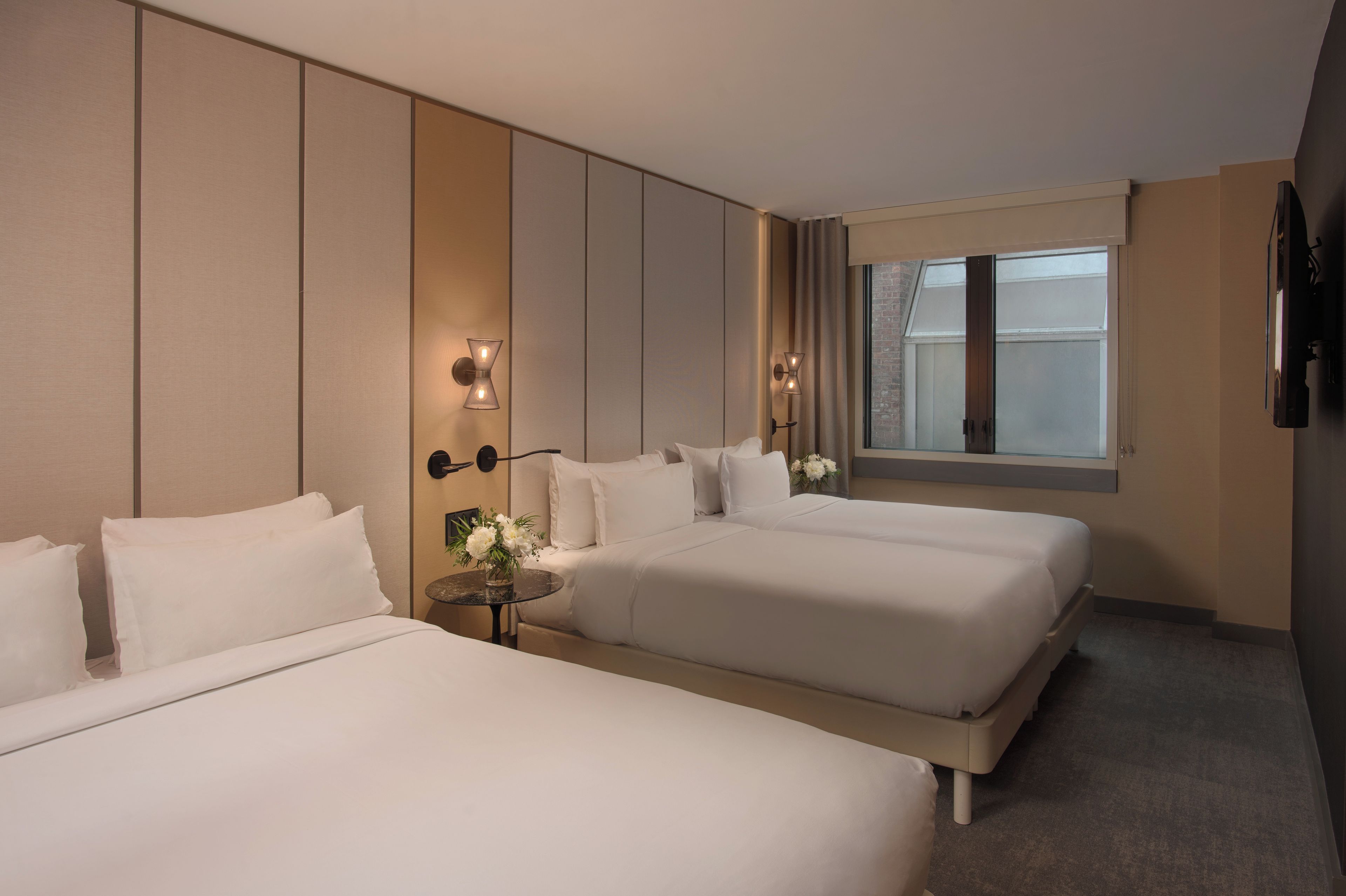 A hotel room with two beds, a small table with a vase of flowers, and a window with curtains.