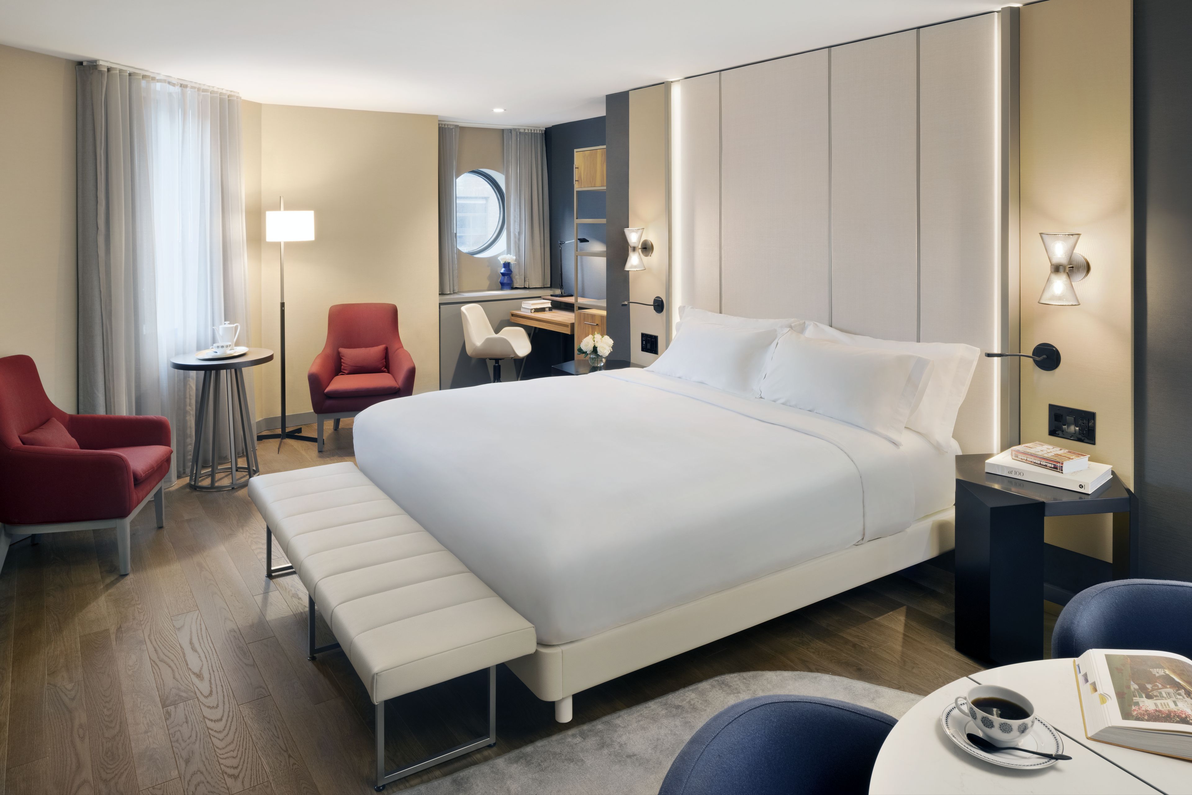 Une chambre d’hôtel avec un grand lit, un bureau, une chaise, un banc et une petite table sur laquelle se trouvent une tasse et un livre.