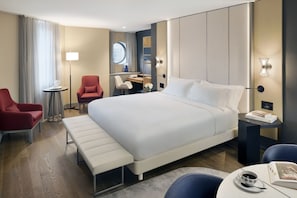 A hotel room with a large bed, a desk, a chair, a bench, and a small table with a cup and book. - NH Collection New York Madison Avenue (New York)