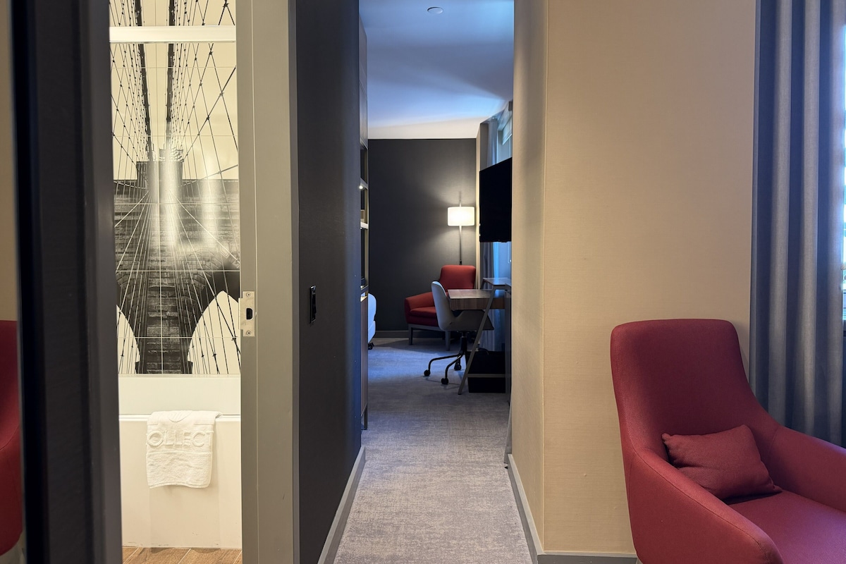 A hotel hallway with a view of a bridge, a red chair, a desk with a lamp, and a white towel hanging on a door.