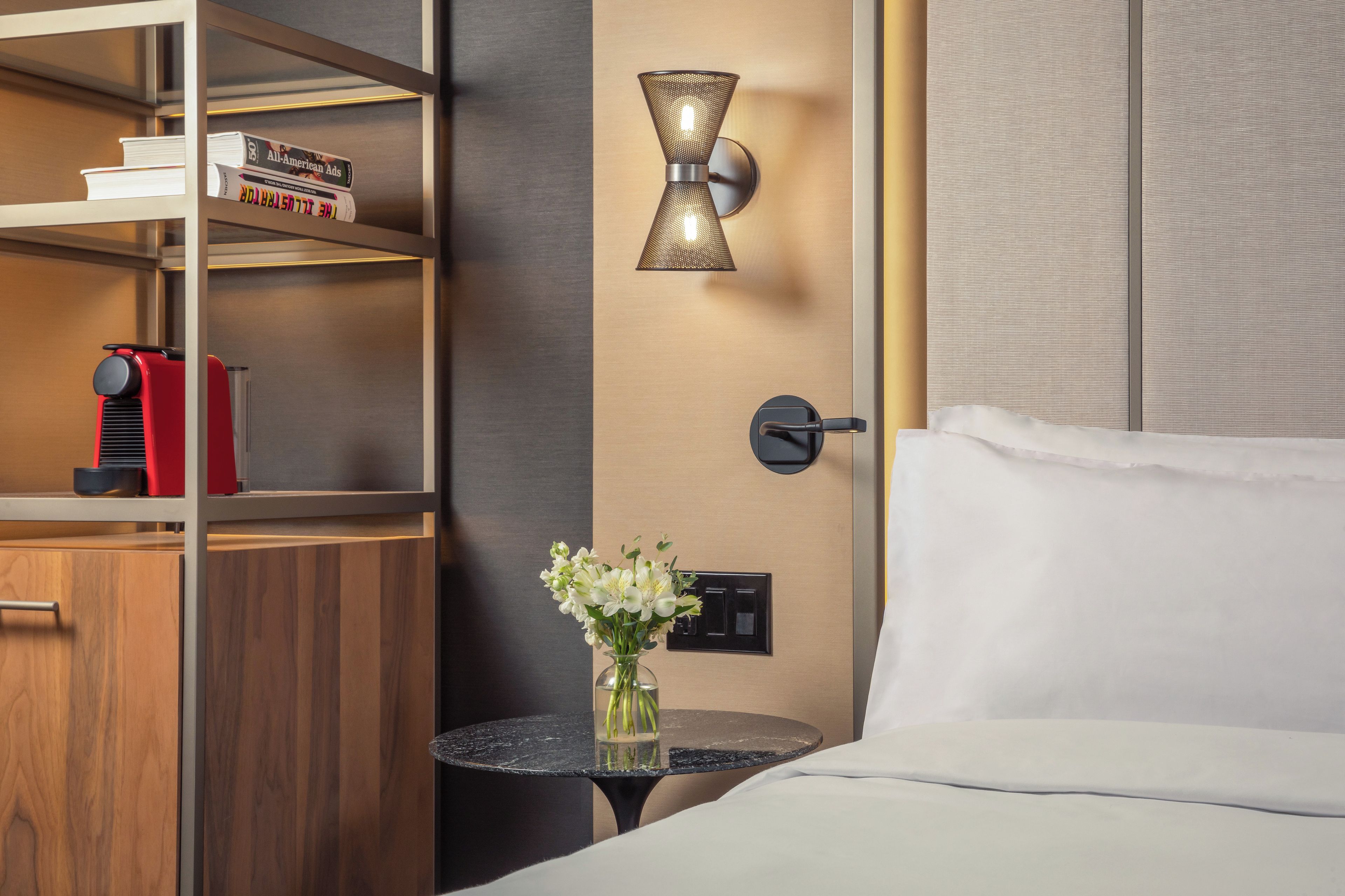 Hotellihuone, jossa on sänky, pieni pöytä maljakko täynnä kukkia, kirjahylly ja seinään kiinnitetty valaisin.