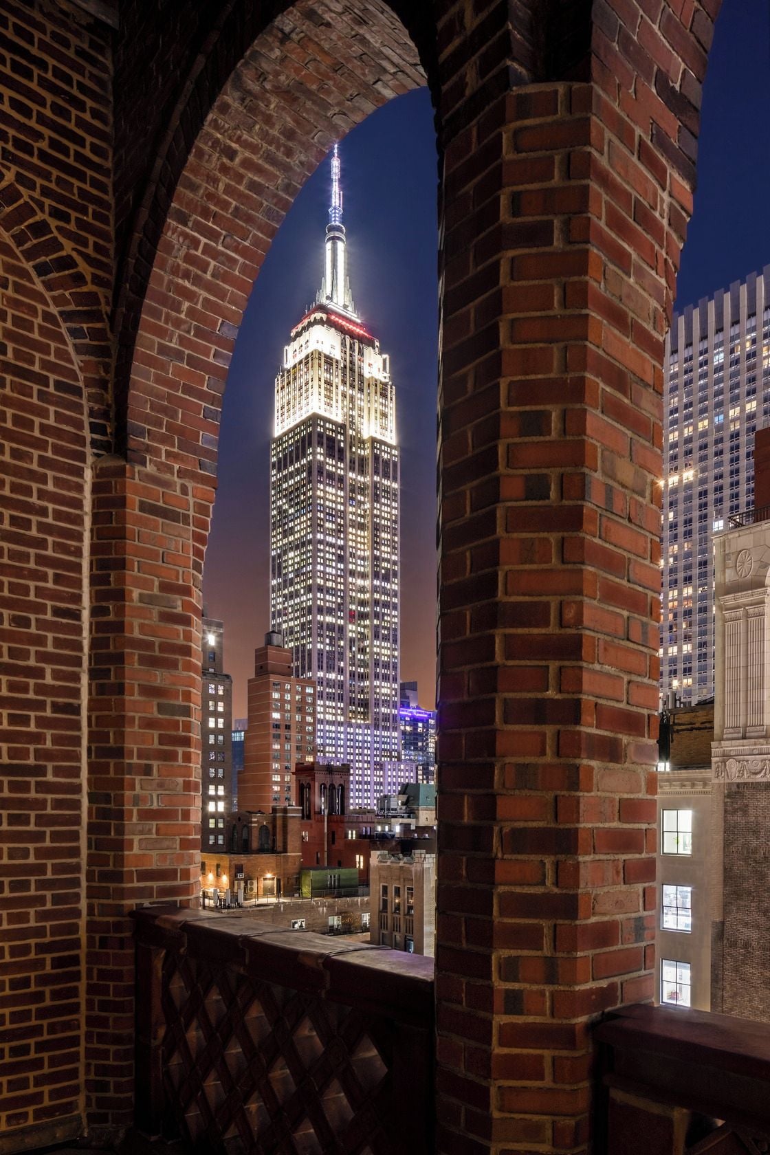 A brick archway framing a view of a tall, illuminated building with a spire, likely a hotel or landmark, with other buildings in the background.