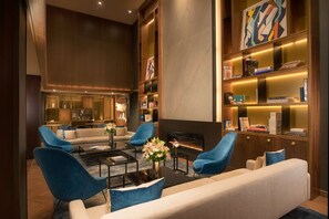 A modern living room with a fireplace, blue armchairs, a sofa, and a bookshelf with books and decorative items. - NH Collection New York Madison Avenue (New York)
